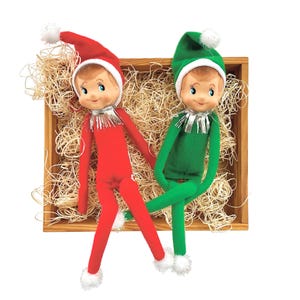 May include: Two Christmas elf dolls in a wooden box filled with straw. One elf wears a red outfit and hat with a white pom-pom, the other wears green. Both have white trim, silver collars, and white fluffy feet.
