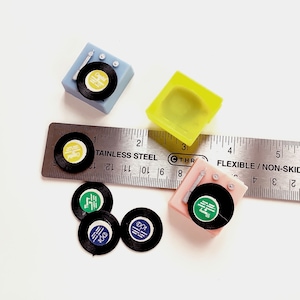 May include: A set of miniature record players, including a blue, pink, and yellow model. Each player has a small record on top with a black and white design. The set is displayed on a ruler with the words "Stainless Steel" and "Flexible/Non-Skid" printed on it.