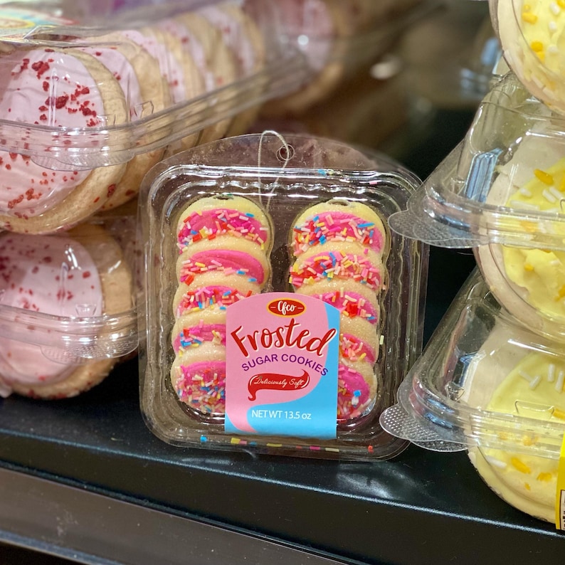 May include: Packaged frosted sugar cookies with pink frosting and colorful sprinkles. The label reads "Frosted Sugar Cookies" and "NET WT 13.5 oz". Other packages of cookies with pink and yellow frosting are visible.