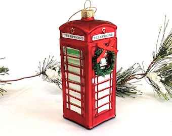 May include: Red glass Christmas ornament shaped like a traditional British telephone booth with a green wreath on the door. The ornament has the word "TELEPHONE" printed on both sides.