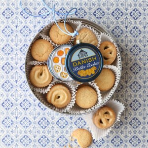Puede incluir: Una lata redonda llena de galletas danesas de mantequilla. La lata tiene una etiqueta azul y blanca que dice "Danish Butter Cookies".