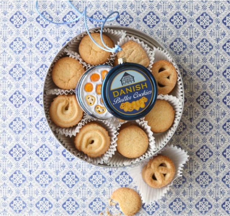 Puede incluir: Una lata redonda llena de galletas danesas de mantequilla. La lata tiene una etiqueta azul y blanca que dice "Danish Butter Cookies".