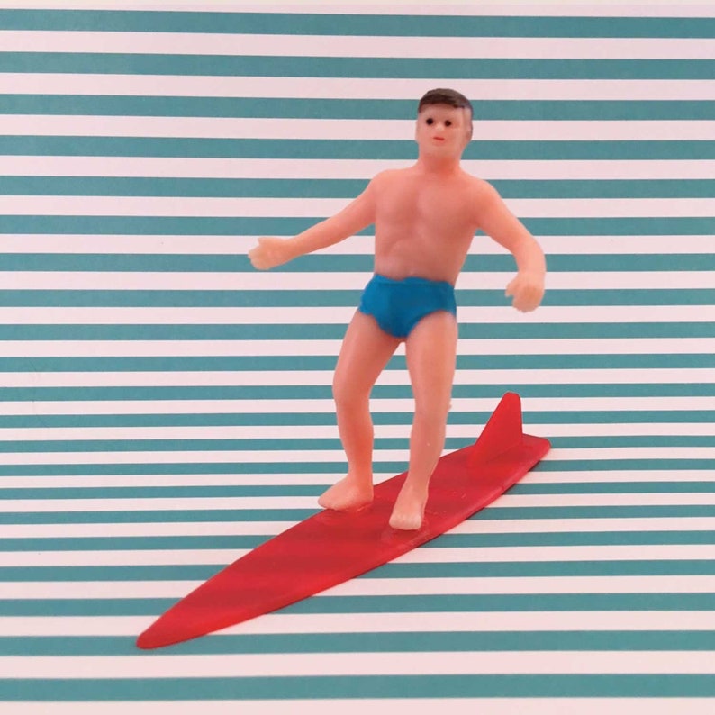May include: A red surfboard with a fin and a figurine of a person wearing blue swim trunks standing on top of it.
