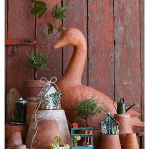 Pode incluir: Uma est&aacute;tua de pato de terracota &eacute; o ponto focal desta exibi&ccedil;&atilde;o com tema de jardim. Estufas em miniatura, um banco de vasos e um regador verde s&atilde;o dispostos com plantas em vasos e vasos de terracota contra um fundo de madeira vermelha.