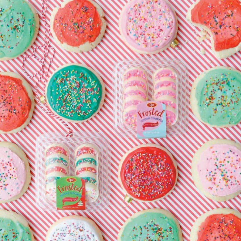 May include: Overhead shot of frosted sugar cookies with colorful sprinkles. Some cookies have red, pink, and green frosting. Several cookies are arranged around two clear plastic containers of frosted cookies. The background is a red and white striped pattern.