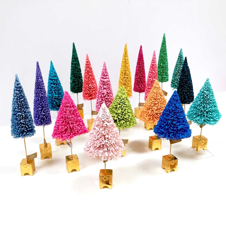 May include: A collection of miniature bottle brush trees in a variety of vibrant colors including pink, blue, green, yellow, and purple. Each tree is mounted on a small, square, metallic gold base and features a thin gold stem.