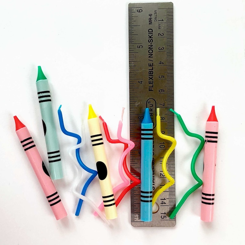 May include: A set of colorful birthday candles with a variety of designs, including a blue candle with a white stripe, a yellow candle with a black dot, and a pink candle with a white stripe. The candles are arranged on a white surface with a ruler in the background.