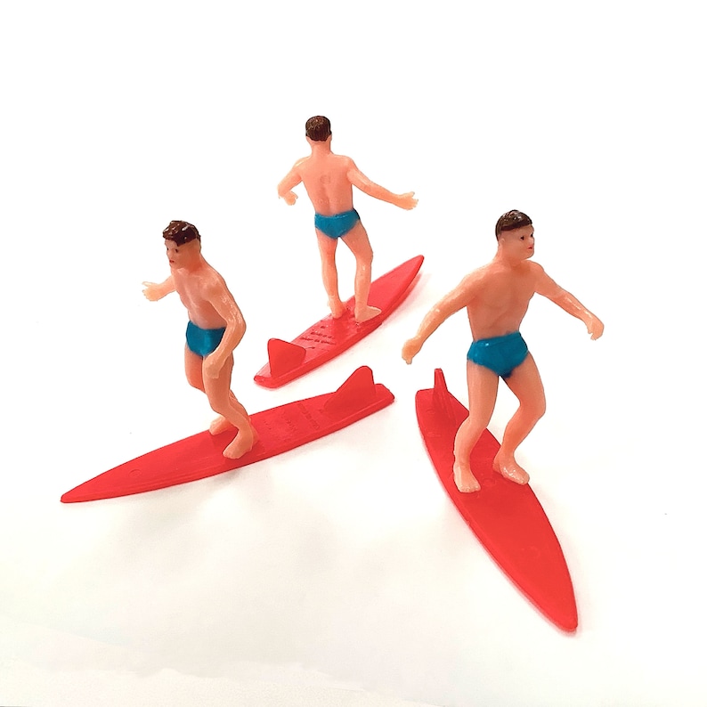 May include: Three red plastic surfboards with three plastic figures standing on them. The figures are wearing blue swim trunks and are in various surfing poses.