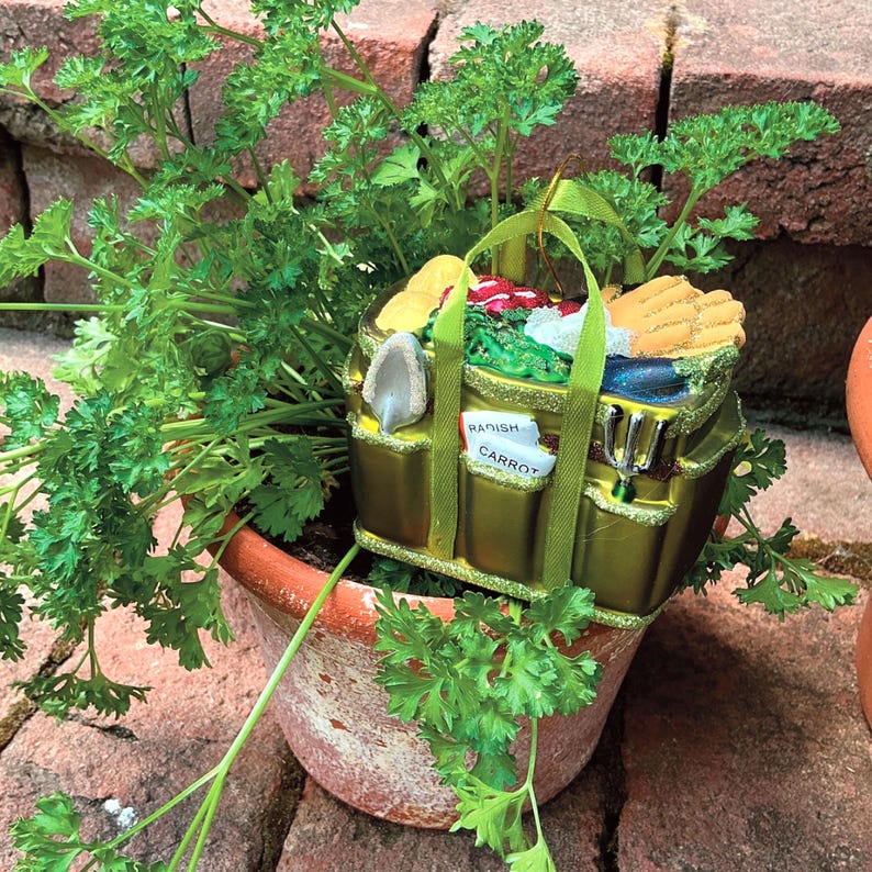 Pode incluir: Um enfeite decorativo com tema de jardim em um vaso de terracota com salsa. O enfeite &eacute; uma sacola dourada cheia de vegetais em miniatura, ferramentas de jardinagem e as palavras "Rabanete" e "Cenoura" em etiquetas.