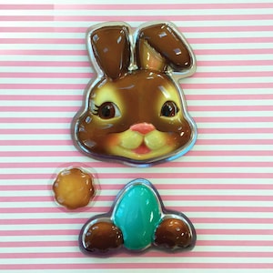 May include: A metal mould for making chocolate Easter bunny shapes. The mould features a bunny face with brown ears and a light blue egg in its paws.