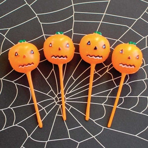 May include: Four orange plastic pumpkin picks, each featuring a carved jack-o'-lantern face with triangular eyes, a small nose, and a jagged mouth. Each pumpkin has a small green stem on top and is attached to a long, thin orange plastic stick.
