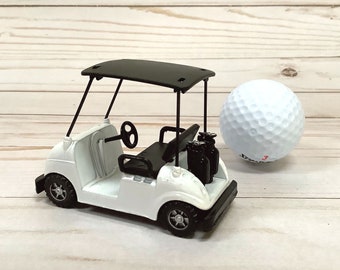May include: A white toy golf cart with a black roof and black wheels. The cart has a black golf bag with golf clubs in the back. A white golf ball is sitting next to the cart.