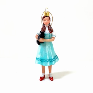 May include: A decorative ornament depicting a girl in a light blue dress, white blouse, and red shoes, holding a black dog. The ornament has a gold halo and a gold loop for hanging. The girl has brown hair in braids.