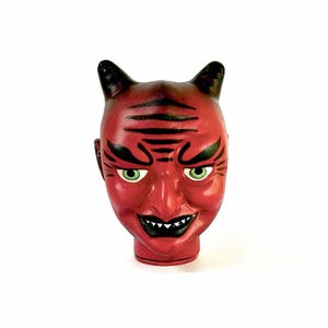 Könnte beinhalten: Eine rote Teufelskopf-Figur mit schwarz bemalten Hörnern, Augenbrauen und Stirnfalten. Das Gesicht hat hellgrüne Augen, eine spitze Nase und einen breiten Mund mit sichtbaren weißen Zähnen. Die Basis des Kopfes ist zylindrisch geformt.