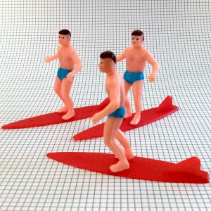 May include: Three plastic figurines of people surfing on red surfboards. The figures are wearing blue swim trunks and have brown hair.