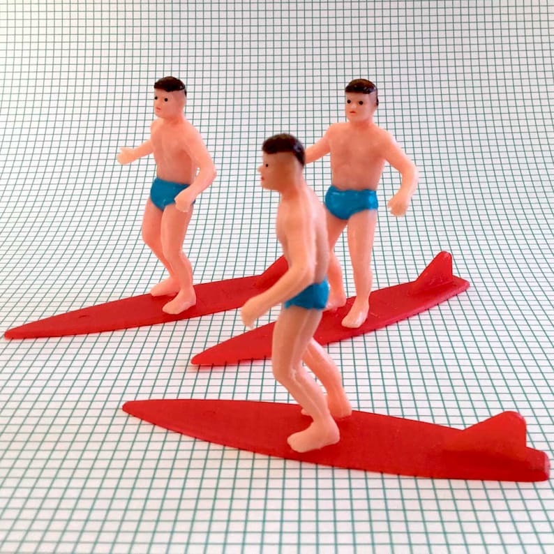 May include: Three plastic figurines of people surfing on red surfboards. The figures are wearing blue swim trunks and have brown hair.