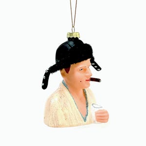 May include: A novelty Christmas ornament depicting a man wearing a black fur hat and smoking a cigar. The ornament is painted with realistic skin tones and details, and is holding a glass. The ornament is suspended by a gold string.