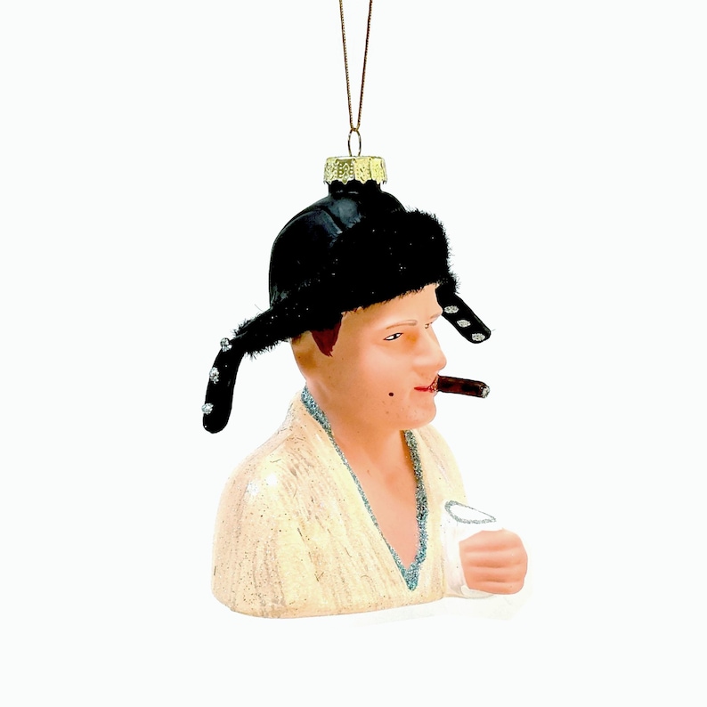 May include: A novelty Christmas ornament depicting a man wearing a black fur hat and smoking a cigar. The ornament is painted with realistic skin tones and details, and is holding a glass. The ornament is suspended by a gold string.
