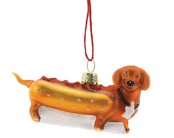 May include: A novelty Christmas ornament shaped like a dachshund hot dog. The ornament is orange with red accents, a black collar, and a white bun. It hangs from a red string and has a gold-colored metal cap.