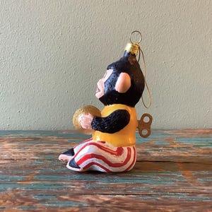 K&ouml;nnte beinhalten: Ein aufziehbares Affen-Weihnachtsornament mit schwarzem Fellmantel, gelbem Hemd und rot-wei&szlig; gestreiften Hosen. Der Affe h&auml;lt einen goldenen Ball in seiner rechten Hand.