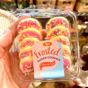 May include: A clear plastic container holds frosted sugar cookies. The cookies have a yellow base, pink frosting, and colorful sprinkles. The label reads "Frosted Sugar Cookies" and "Deliciously Soft". The net weight is 13.5 oz.