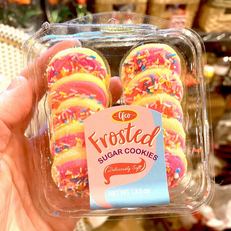 May include: A clear plastic container holds frosted sugar cookies. The cookies have a yellow base, pink frosting, and colorful sprinkles. The label reads "Frosted Sugar Cookies" and "Deliciously Soft". The net weight is 13.5 oz.