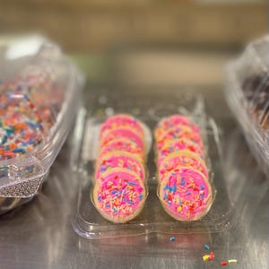 May include: A clear plastic container holds a row of frosted sugar cookies. Each round cookie has a pink frosting and is topped with colorful sprinkles. Another container to the left has cookies with colorful sprinkles.