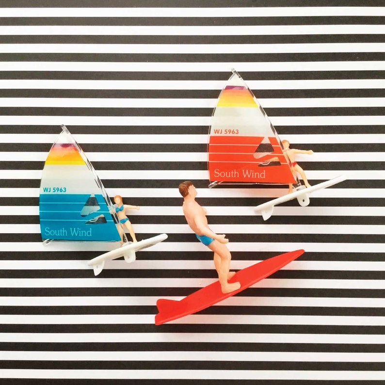 May include: Two miniature sailboats with the text "WJ 5963 South Wind" on them. One sailboat is red and white, the other is blue and white. A miniature person is surfing on a red surfboard.
