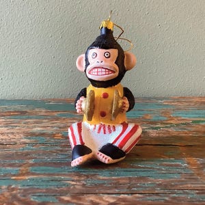 K&ouml;nnte beinhalten: Ein gl&auml;serner Weihnachtsbaumschmuck mit einem Affen in einem gelben und roten Outfit, der Becken spielt. Der Affe hat ein schwarzes Gesicht und ein breites Grinsen.