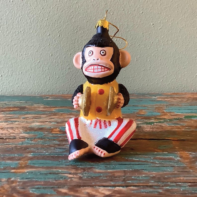 K&ouml;nnte beinhalten: Ein gl&auml;serner Weihnachtsbaumschmuck mit einem Affen in einem gelben und roten Outfit, der Becken spielt. Der Affe hat ein schwarzes Gesicht und ein breites Grinsen.