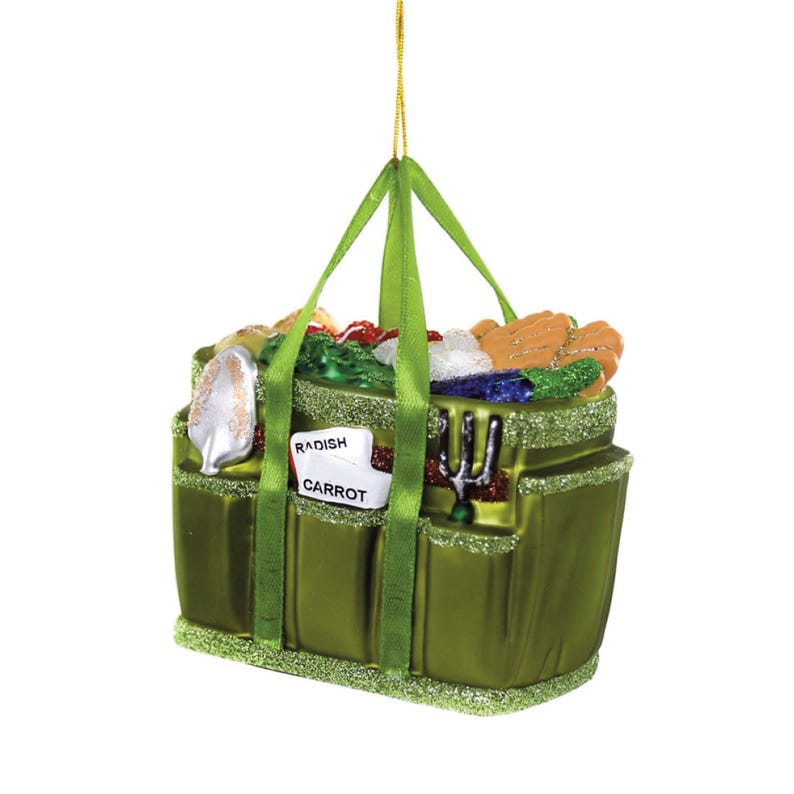May include: A green glass ornament shaped like a gardening tote bag. The tote is filled with colorful vegetables and gardening tools. A tag reads "Radish Carrot". The ornament has green handles and glitter accents.