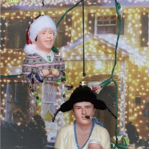 May include: Two Christmas ornaments depicting characters from a holiday movie. One ornament features a figure wearing a Santa hat and a sweater, while the other has a fur hat and a cigar. The background is a blurred image of a house with Christmas lights.
