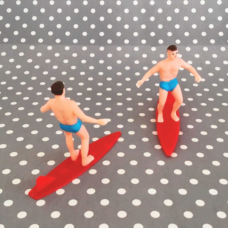 May include: Two miniature plastic figures of surfers riding red surfboards. The figures are wearing blue swim trunks and have brown hair. The figures are posed as if they are surfing on waves.