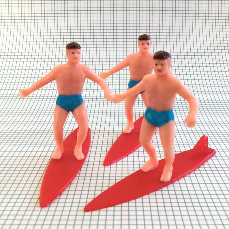 May include: Three plastic figurines of people surfing on red surfboards. The figures are wearing blue swim trunks and have pink skin.