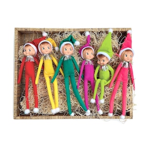 May include: Six colorful elf dolls in a wooden crate. Each doll wears a different colored outfit: red, yellow, green, purple, lime green, and pink. They have white pom-poms on their hats and silver tinsel collars.