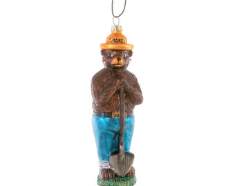 Può includere: Un ornamento di vetro di Smokey Bear, la mascotte del Servizio Forestale degli Stati Uniti, che indossa un cappello da ranger e tiene una pala. L'ornamento è marrone, blu e arancione.