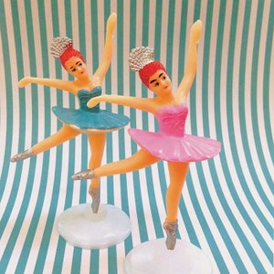 Könnte beinhalten: Zwei Plastik-Ballerina-Figuren in Tanzpose. Eine Ballerina trägt einen rosa Tüllrock, die andere einen blauen Tüllrock. Beide haben rotes Haar, silberne Diademe und silberne Ballettschuhe. Sie stehen auf weißen Sockeln vor einem gestreiften Hintergrund.