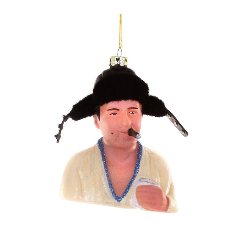 May include: A novelty Christmas ornament featuring a figure wearing a black fur hat and smoking a cigar. The ornament has a cream-colored shirt with blue trim and is holding a white cloth. The ornament is suspended by a gold string.