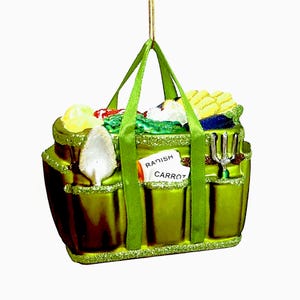 Pode incluir: Um enfeite de saco de jardim verde com um fio dourado para pendurar. O saco &eacute; preenchido com vegetais em miniatura e ferramentas de jardinagem. Uma etiqueta diz "Rabanete Cenoura". O enfeite tem uma borda brilhante e al&ccedil;as verdes.