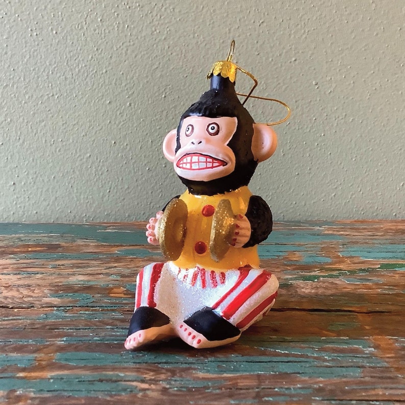 K&ouml;nnte beinhalten: Ein schwarzer und gelber Glas-Anh&auml;nger mit einem Affen, der Becken spielt. Der Affe tr&auml;gt ein rot-wei&szlig; gestreiftes Outfit und hat einen schelmischen Ausdruck.