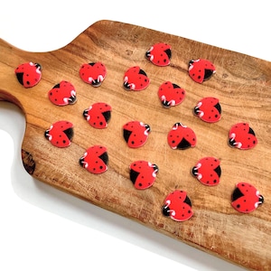 May include: A collection of red ladybug-shaped embellishments with black spots and wings, arranged on a wooden cutting board. The ladybugs are evenly spaced, creating a visually appealing pattern. The wooden board has a natural wood grain.
