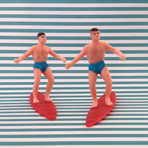 May include: Two plastic figurines of people surfing on red surfboards. The figures are wearing blue swim trunks and have brown hair. The background is a teal and white striped pattern.