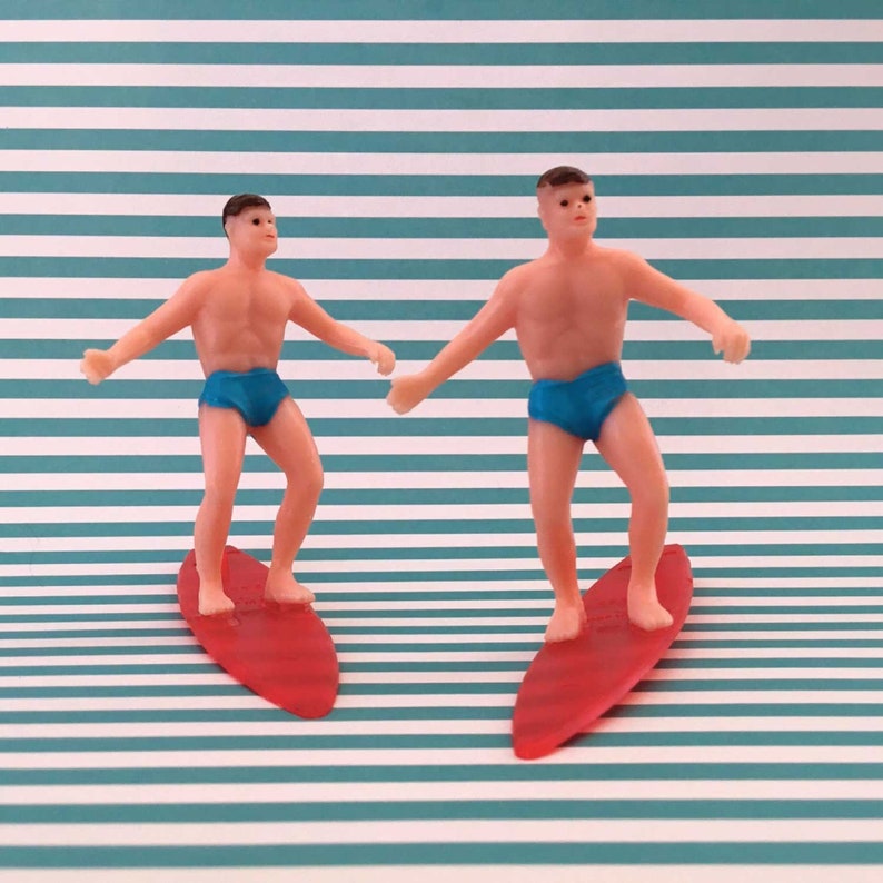 May include: Two plastic figurines of people surfing on red surfboards. The figures are wearing blue swim trunks and have brown hair. The background is a teal and white striped pattern.