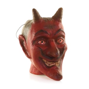 May include: A red devil mask with a sinister grin, pointed horns, and red eyes. The vintage Halloween mask has a wire attached for wearing. The devil's face is painted with dark accents, creating a spooky and detailed appearance.