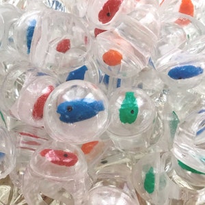 Może przedstawiać: Kolekcja pierścionków z przezroczystego plastiku, z których każdy zawiera małą, jednokolorową figurkę ryby zamkniętą w przezroczystej, sferycznej kopule. Ryby są w żywych odcieniach czerwieni, błękitu, zieleni i pomarańczu, każda z czarnym okiem.