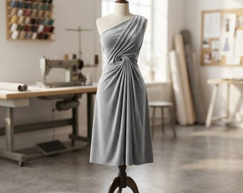 Puede incluir: Un vestido gris claro hasta la rodilla expuesto en un maniquí. El vestido presenta un solo tirante y un distintivo detalle fruncido circular en la cintura, creando pliegues verticales suaves que caen a lo largo de la falda de la prenda.