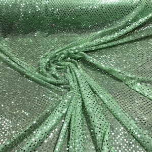 May include: Close-up of a shimmering, mint green fabric covered in small, reflective sequins. The fabric is draped and folded, creating a textured, flowing appearance. The sequins catch the light, adding sparkle.