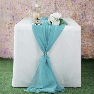 May include: A teal chiffon table runner with a crystal embellishment, placed on a white tablecloth. A white flower bouquet and a decorative birdcage are on the table.