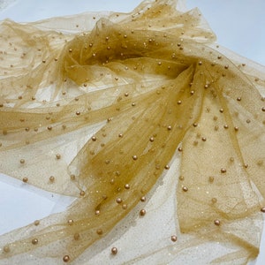 May include: Gold tulle fabric with small gold beads scattered throughout. The fabric is draped and folded, showing the texture and sparkle of the material.