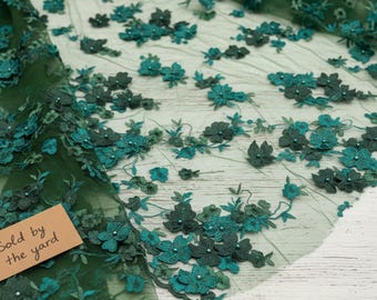May include: A sheer dark green tulle fabric featuring three-dimensional floral embroidery in various shades of green, accented with small beads at the center of each flower. A small tan tag with the text Sold by the yard is attached.
